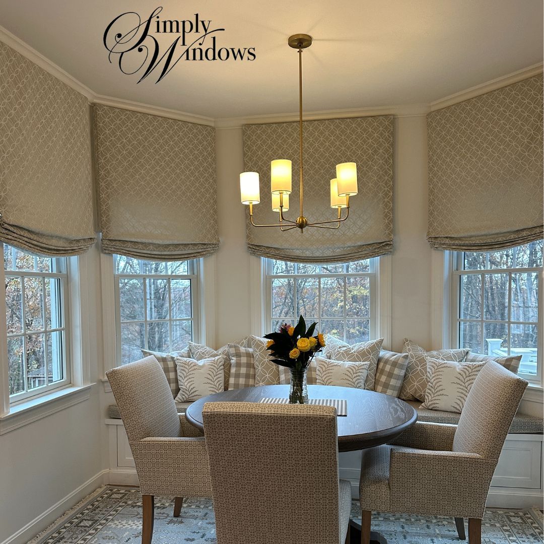 Bright breakfast nook with round table, wicker chairs, bay windows, roman shades, and a floral centerpiece.