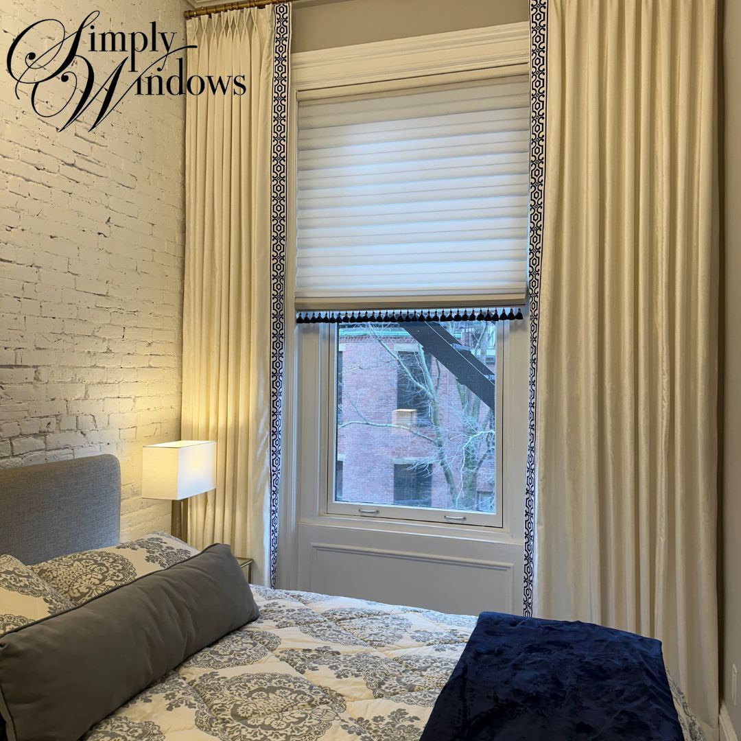 Cozy bedroom with patterned bed, lamp, large window, and cream custom drapery.