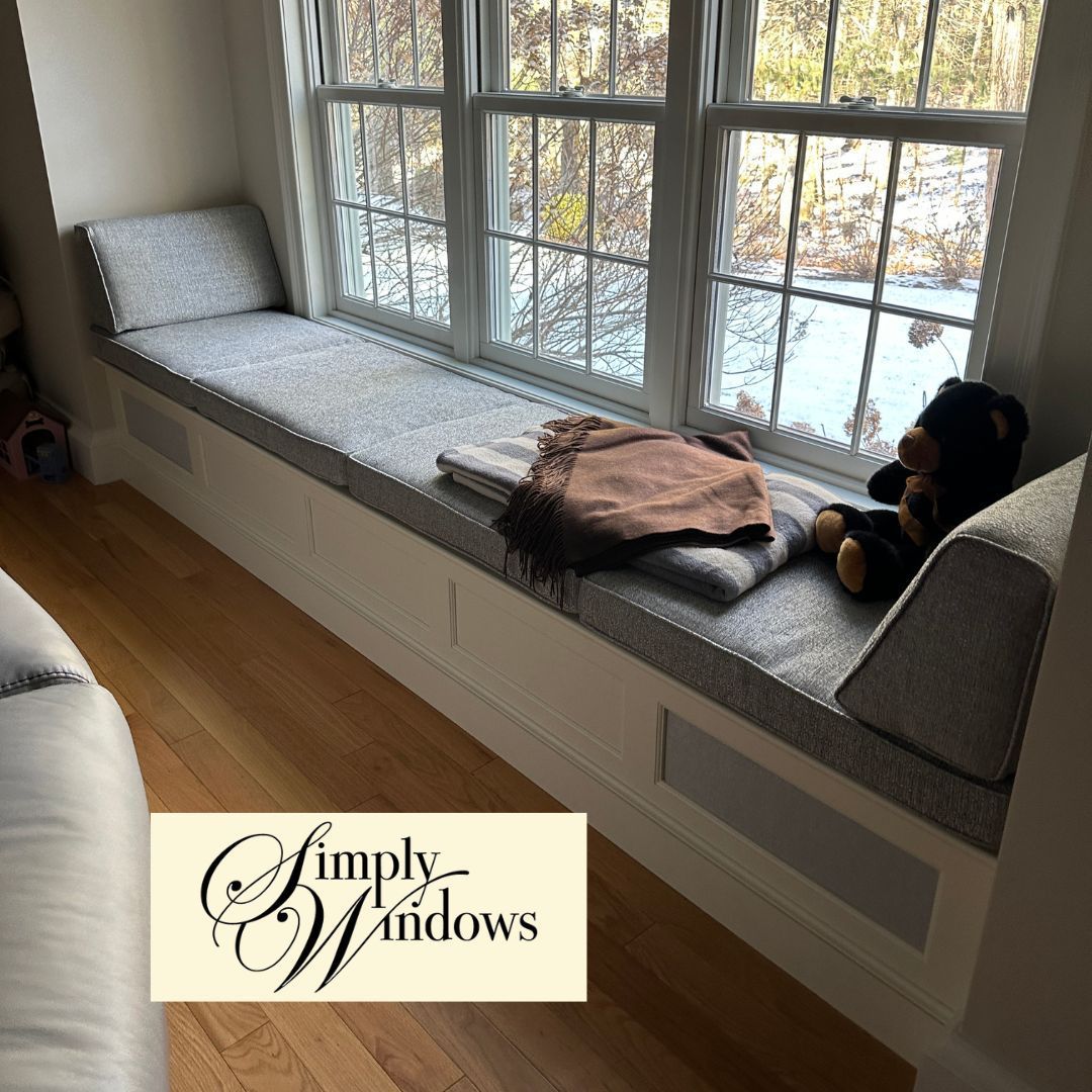 Window seat with gray designer fabric cushions, folded blanket, and teddy bear in a bright room