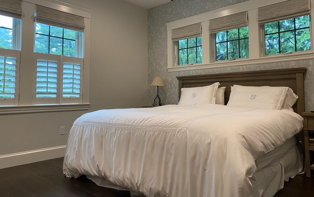Bedroom with a large white bed, dark flooring, and windows with shutters and roman shades on two walls.