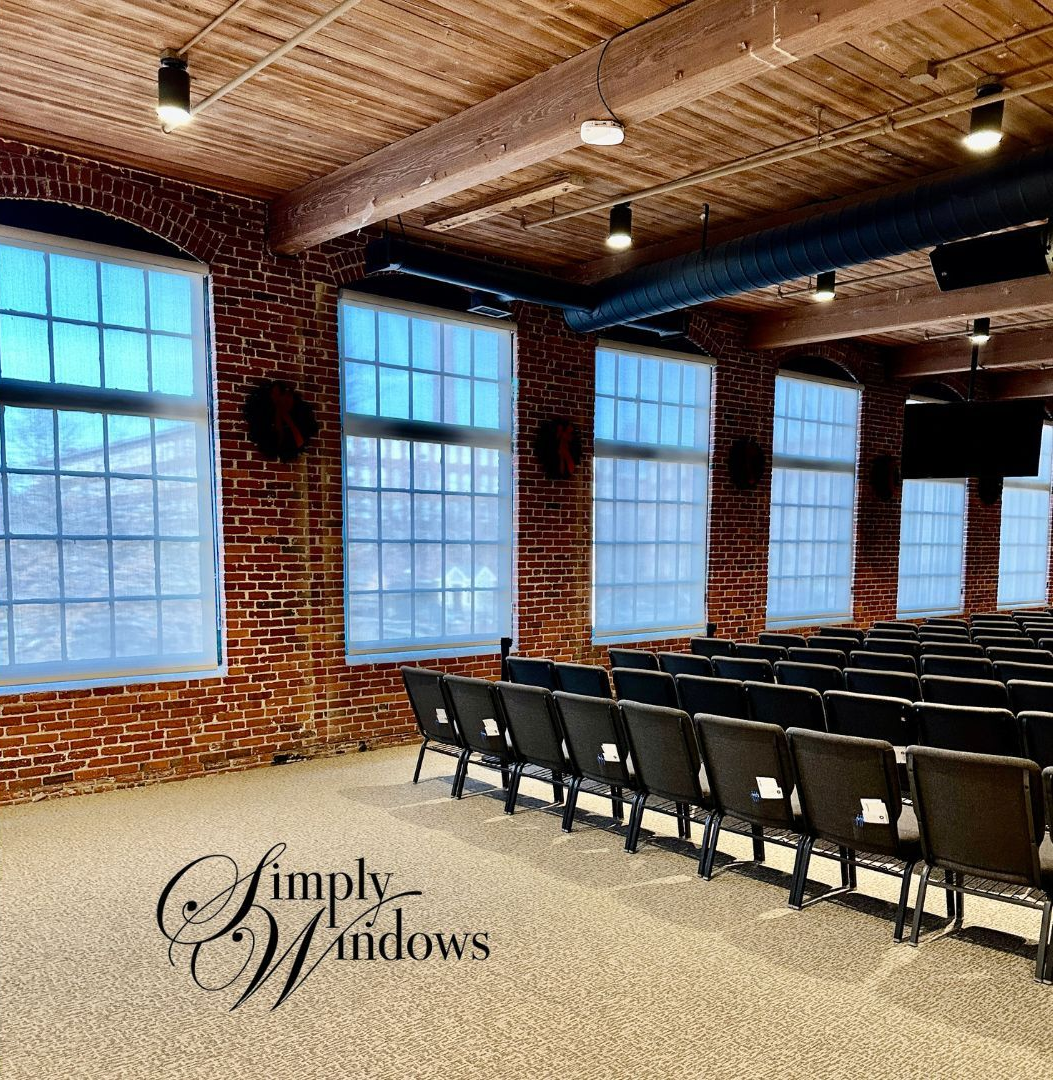 Brick-walled event room with large windows with solar shades, rows of black chairs, and a textured ceiling.