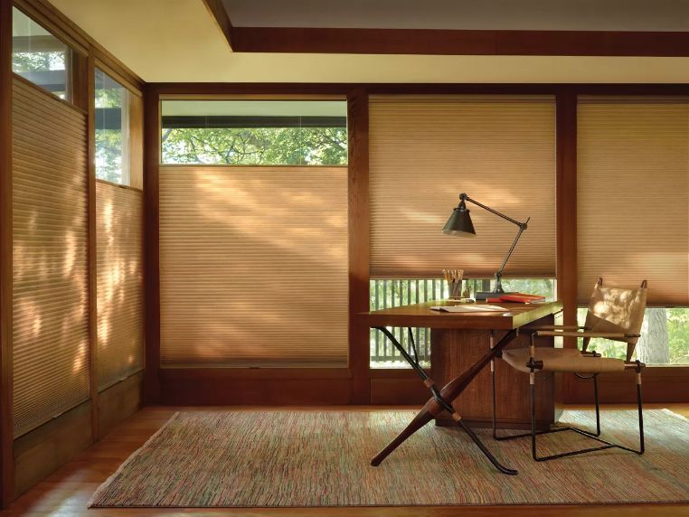 Cozy home office with closed tan blinds. Wooden desk, lamp, chair, and rug with sunlight shining in.