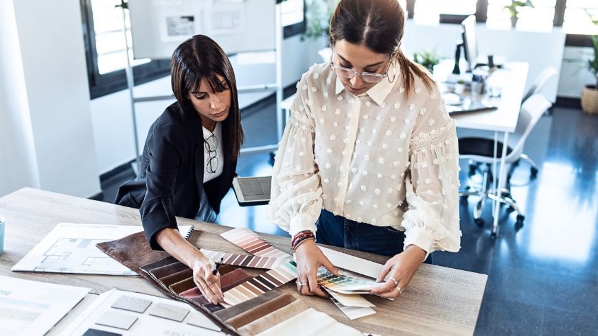 two women are sitting at a table looking at samples of fabric Simply Windows.