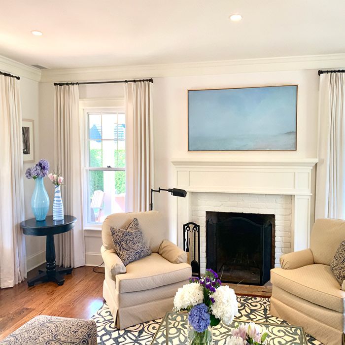 Cozy living room with fireplace, white walls, and beige armchairs. Blue artwork hangs above the mantel.