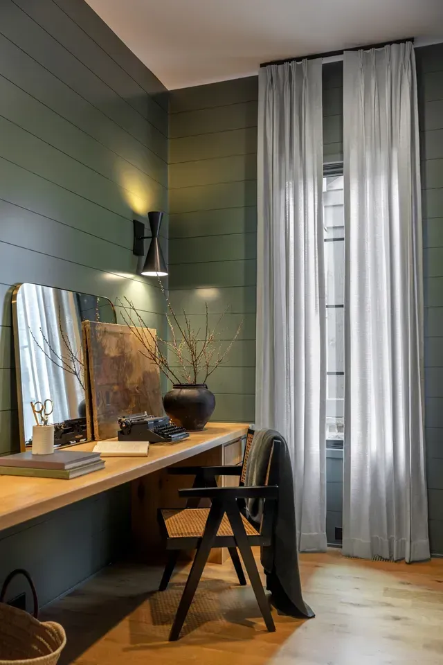 A home office with a wooden desk, a black chair, and large white curtains. Green paneled walls.