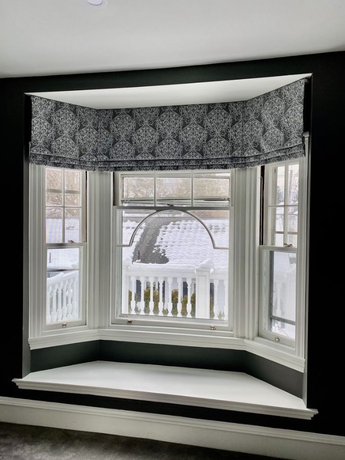 Bay window with dark patterned Roman shade, white trim, and a snowy outdoor view.