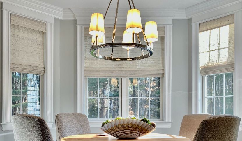 A dining room has three windows behind it with woven wood bamboo roman shades.