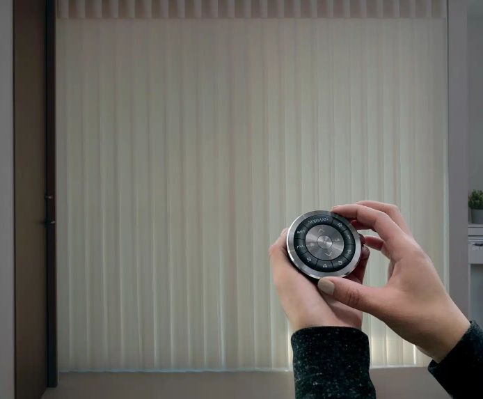 Person holding a circular remote control, likely for vertical blinds. Blinds are beige.