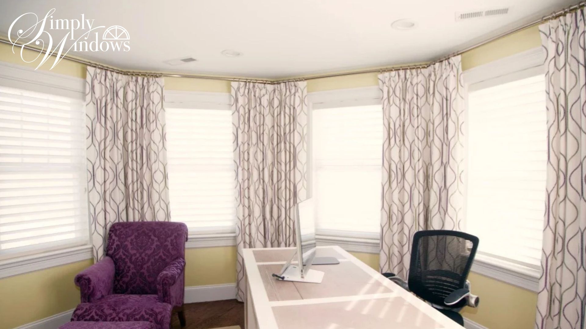 Bright bay-window home office with a white desk, black chair, purple armchair, and patterned curtains.