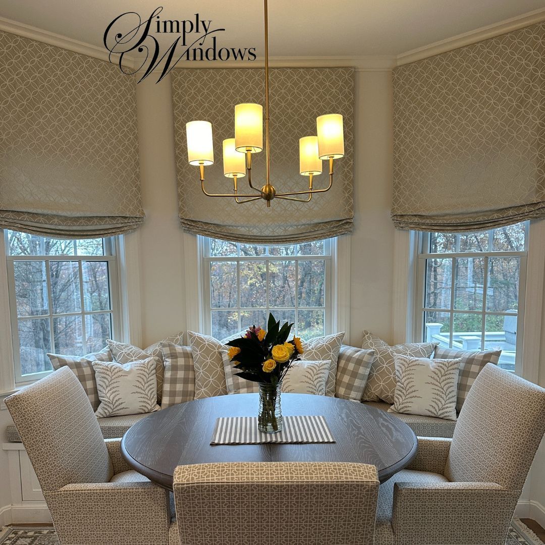 A dining nook with a round table, chairs, built-in window seating, patterned Roman shades, and a gold chandelier.