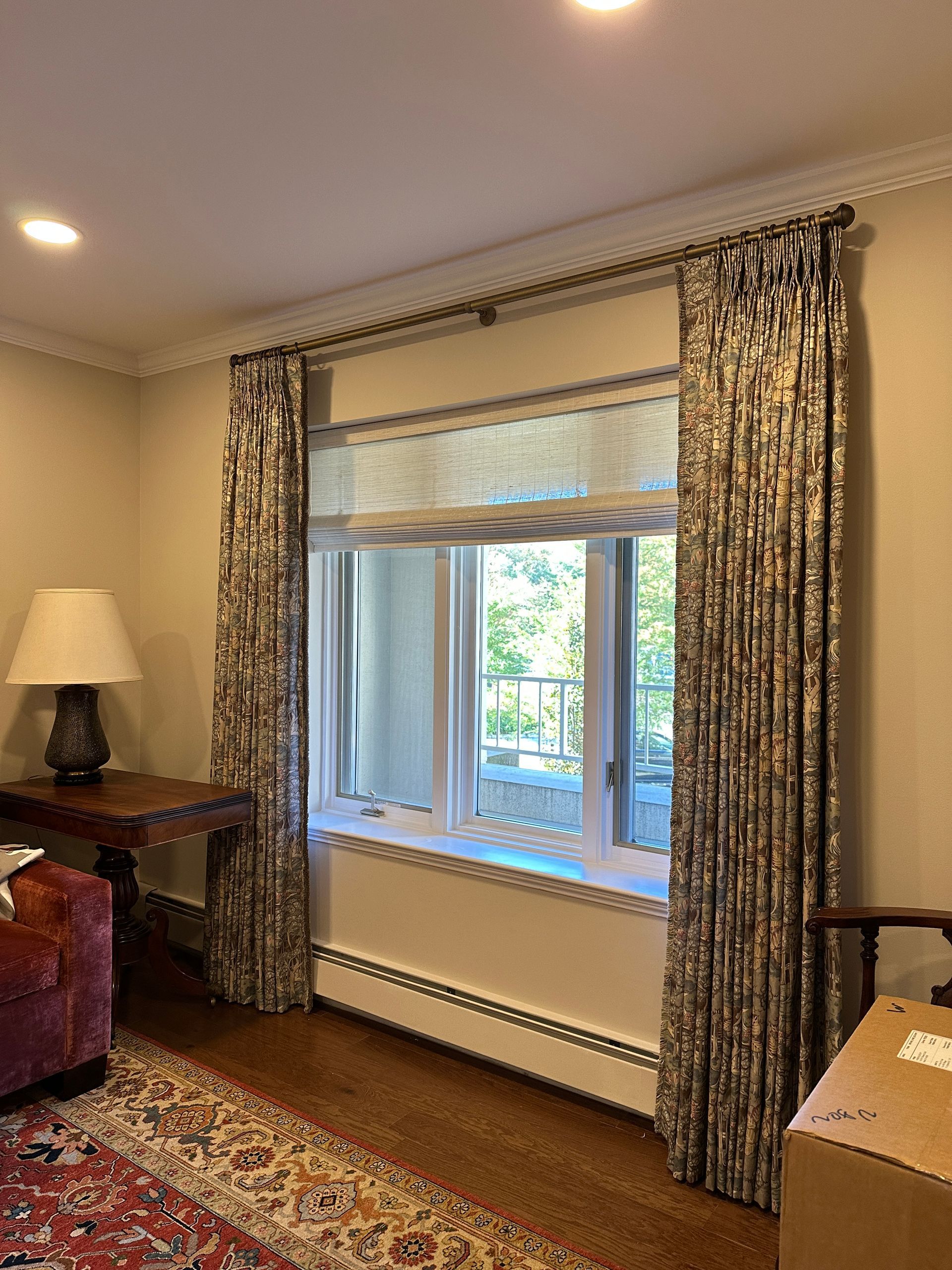 Window with patterned curtains and a Roman shade, near a table and lamp, in a room with hardwood floors.