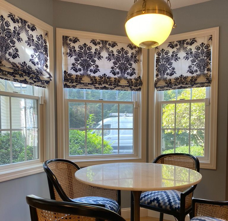 A dining nook with a white round table, black wicker chairs with patterned cushions, and windows with floral Roman shades.