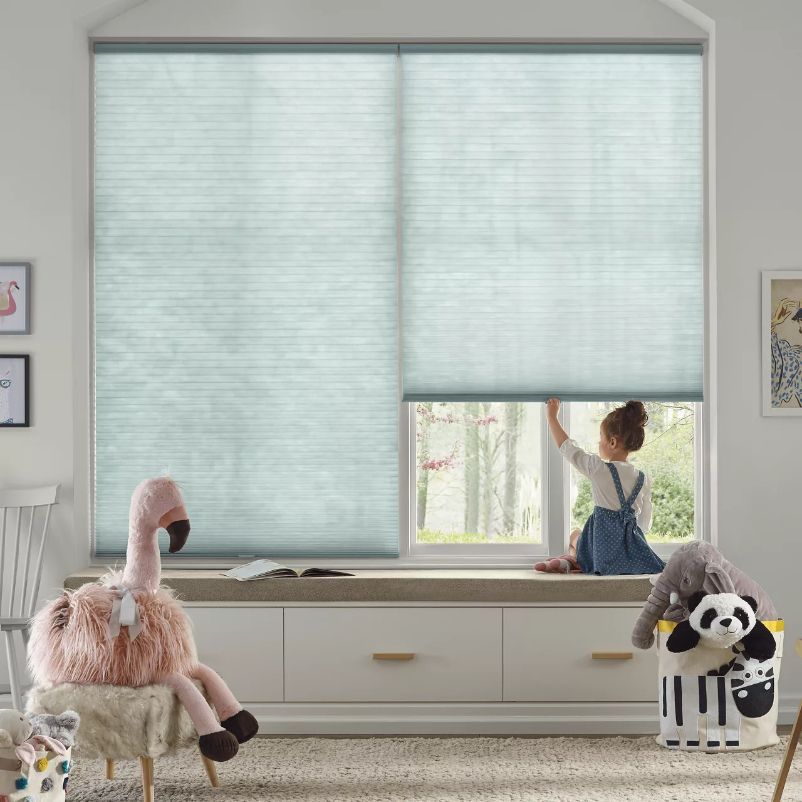 Child looking out window, light blue honeycomb blinds. Window seat with stuffed animals: flamingo, panda.