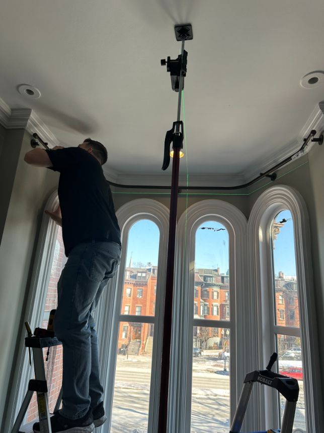 Man on stepladder installing curtain rod near curved windows; laser level.