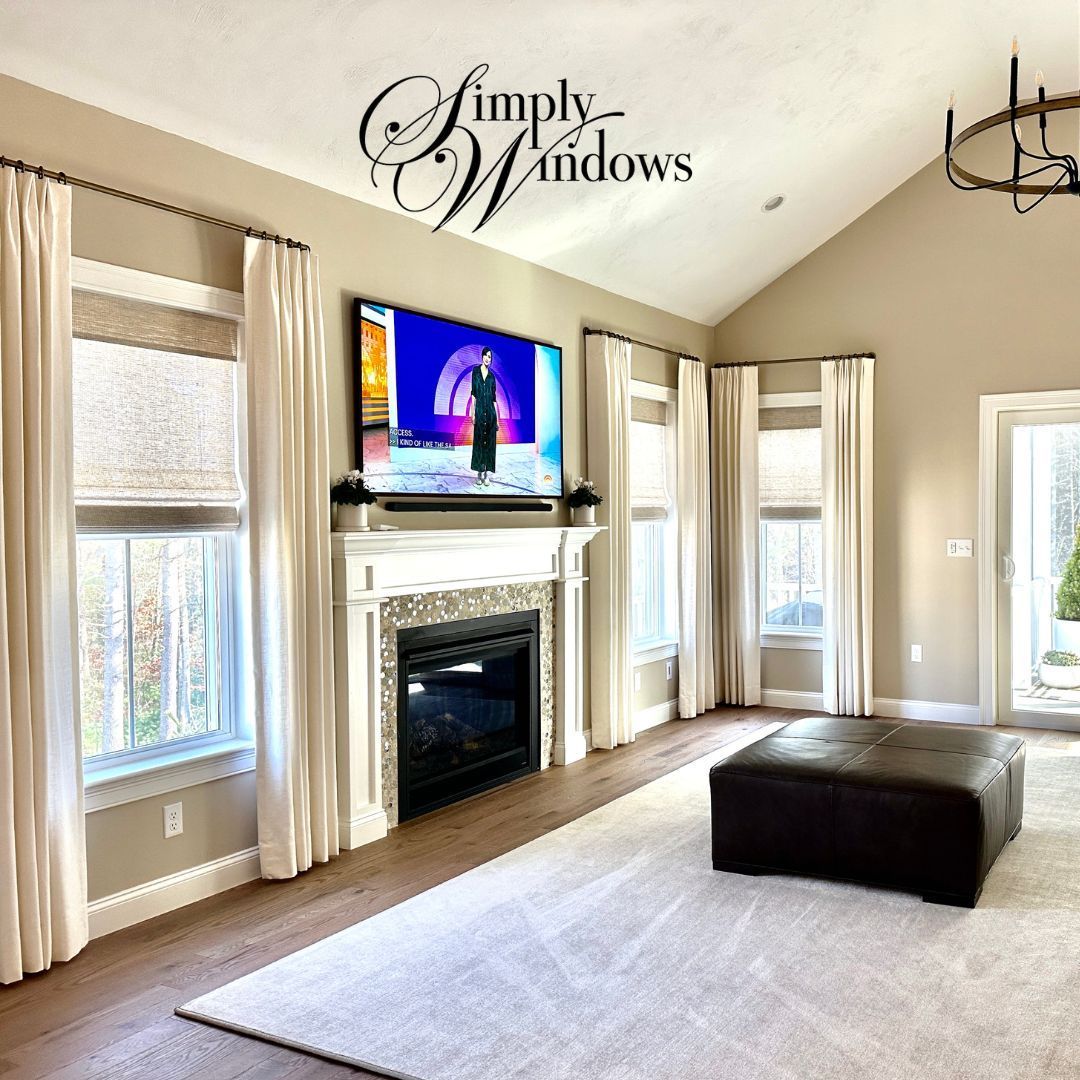 Bright living room with fireplace, TV, large windows with layered window treatments, and a dark ottoman on a light rug