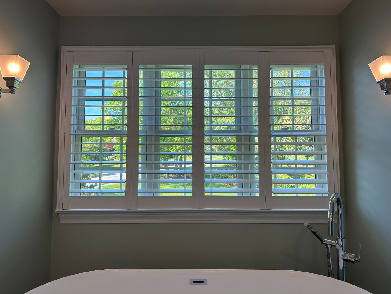 Bathroom with white plantation shutters over a tub and chrome faucet, lit by wall sconces