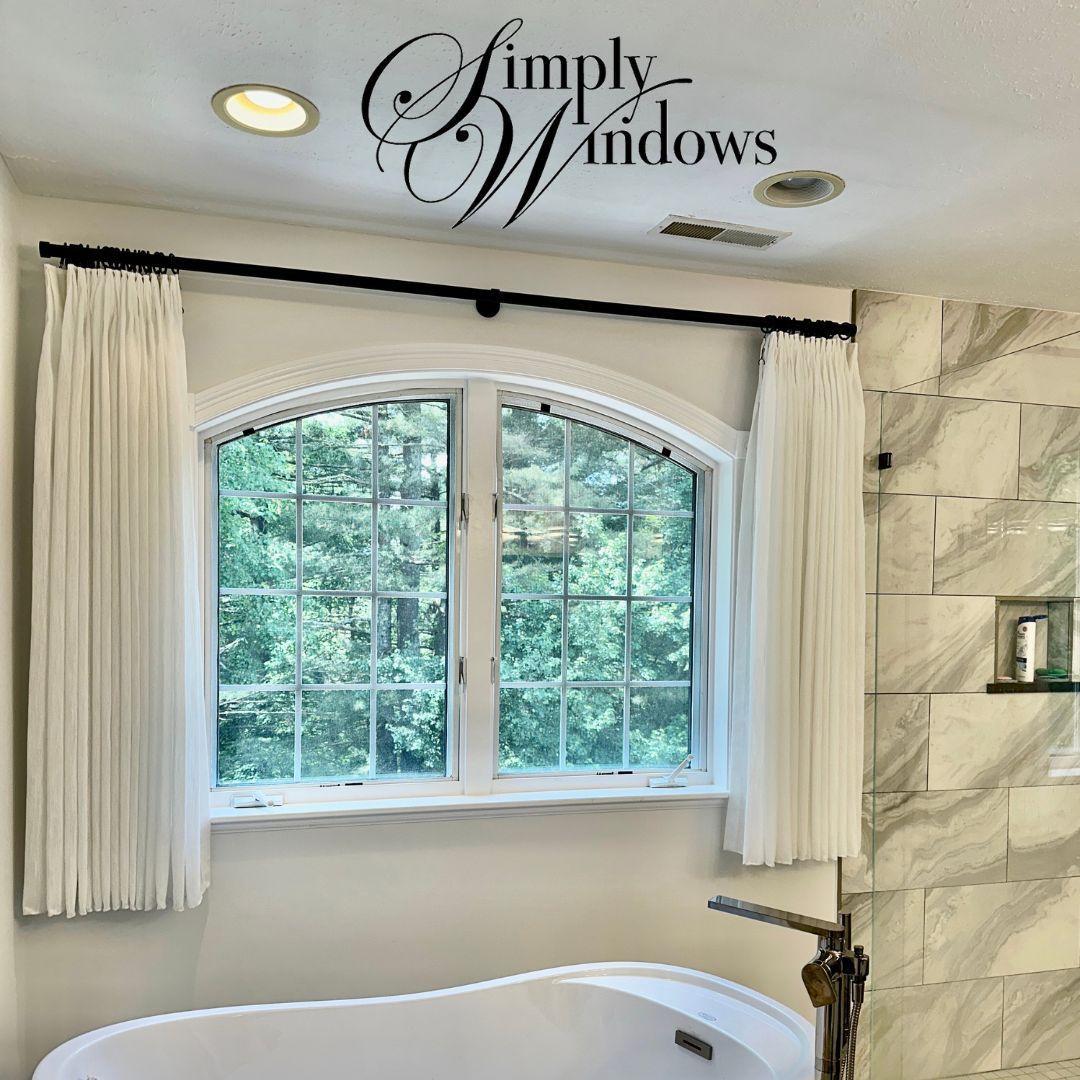 Bathroom with white arched window with custom curtains, beige walls, and a freestanding tub.