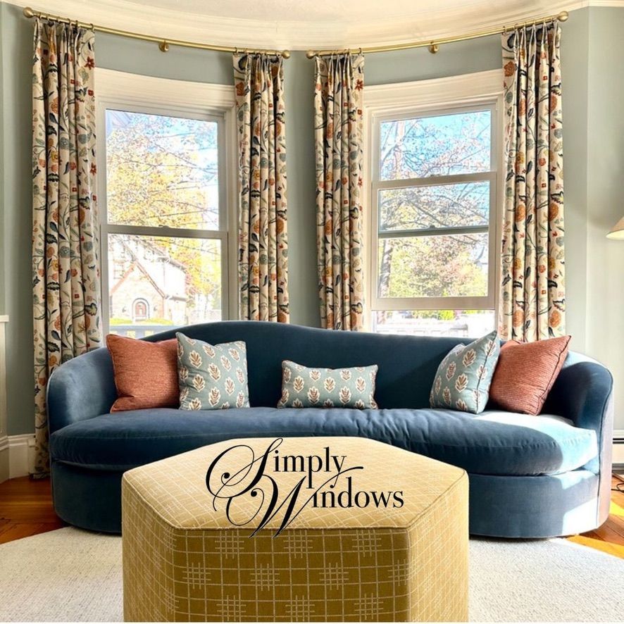 Curved blue sofa in a bright bay window room with patterned drapery and a gold ottoman