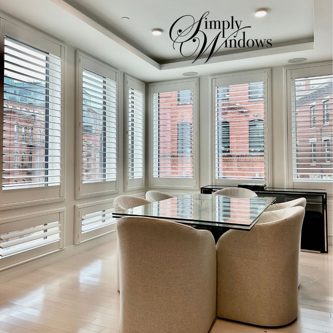 Bright bay-windowed room with a glass table, beige chairs, and white shutters, in a modern dining room.