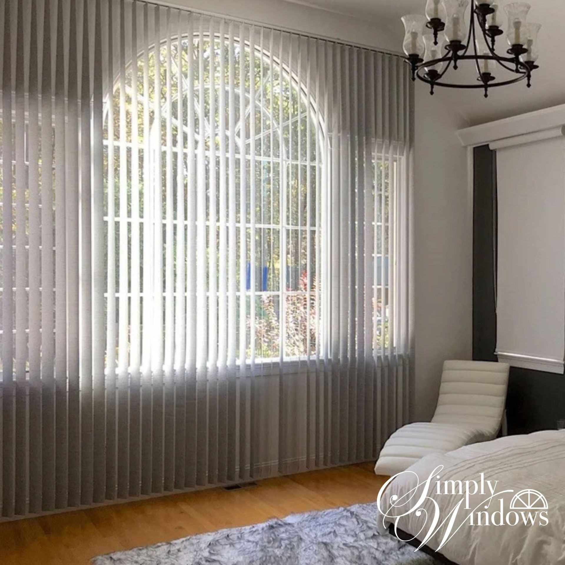 Bright living room with arched window, vertical blinds, wood floor, gray chair, and black chandelier