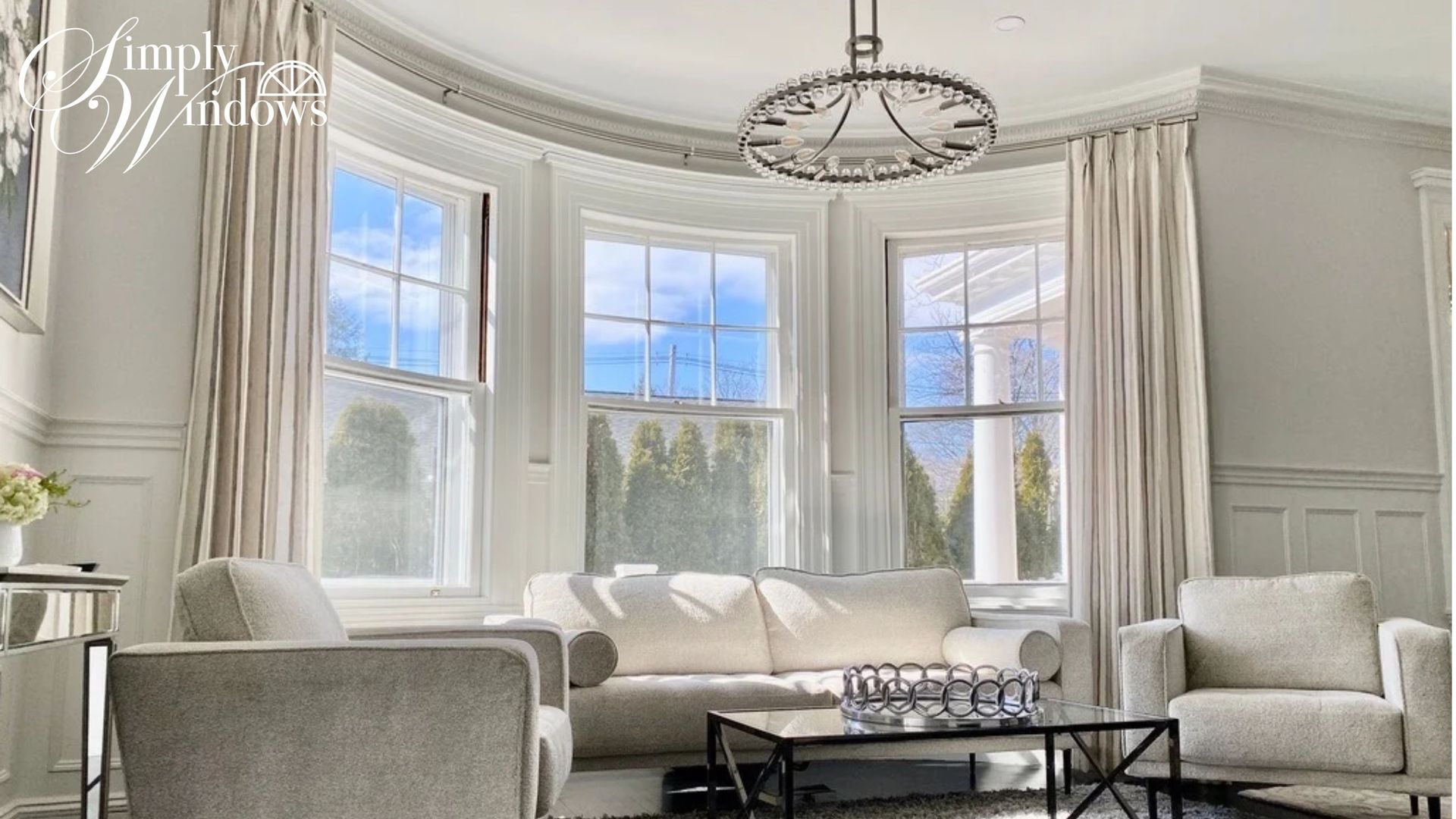 Bright, elegant living room with large bay windows, white seating, custom white drapery, and a chandelier