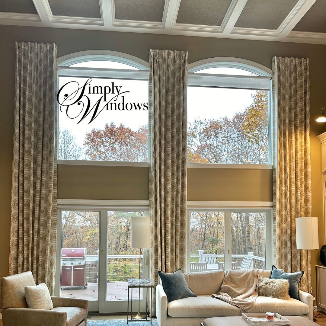 Bright living room with tall arched windows, beige drapery, and a sofa with pillows overlooking autumn trees.