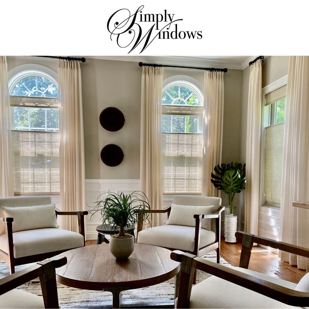 Bright living room with cream chairs, round wood table, tall arched windows with layered window treatments. 