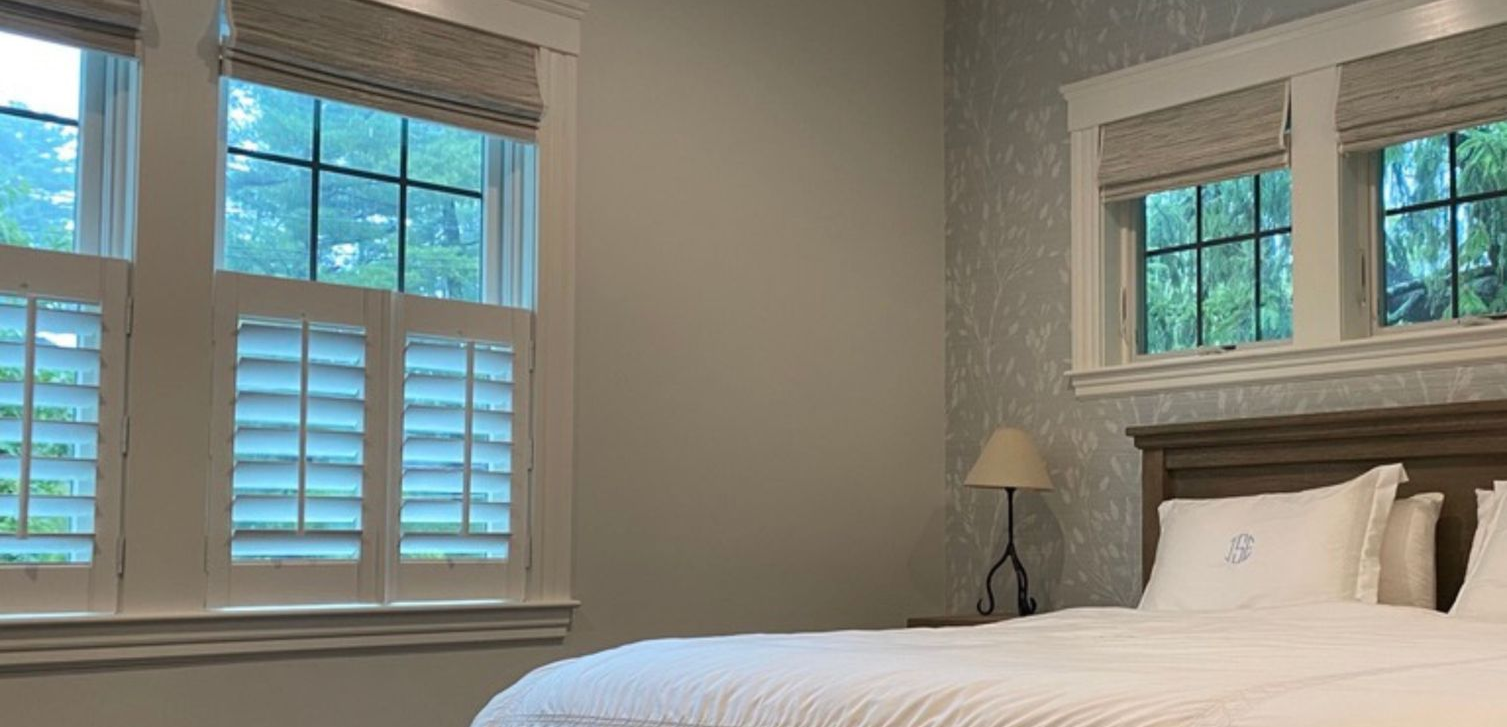 Bright bedroom with a white bed, bedside lamp, and two windows with shutters and blue sky outside