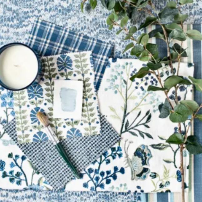 a table topped with a variety of blue and white fabrics and a candle Simply Windows.