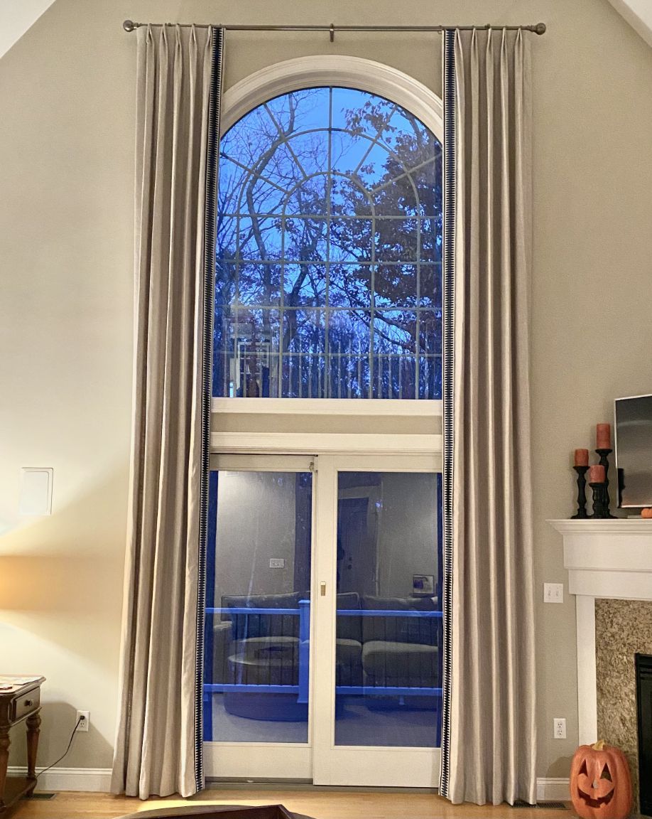 Tall arched window with full-length beige drapes. French doors below. Outside view shows blue sky and trees.