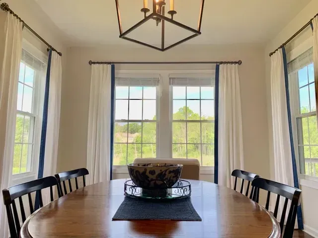 Dining room with large windows, white curtains with blue trim, and wooden table with black chairs.