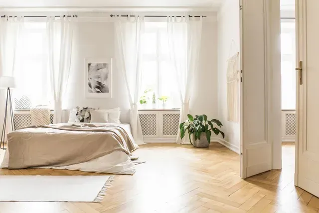 Light-filled bedroom with hardwood floors, a bed with tan bedding, white curtains, and a potted plant.