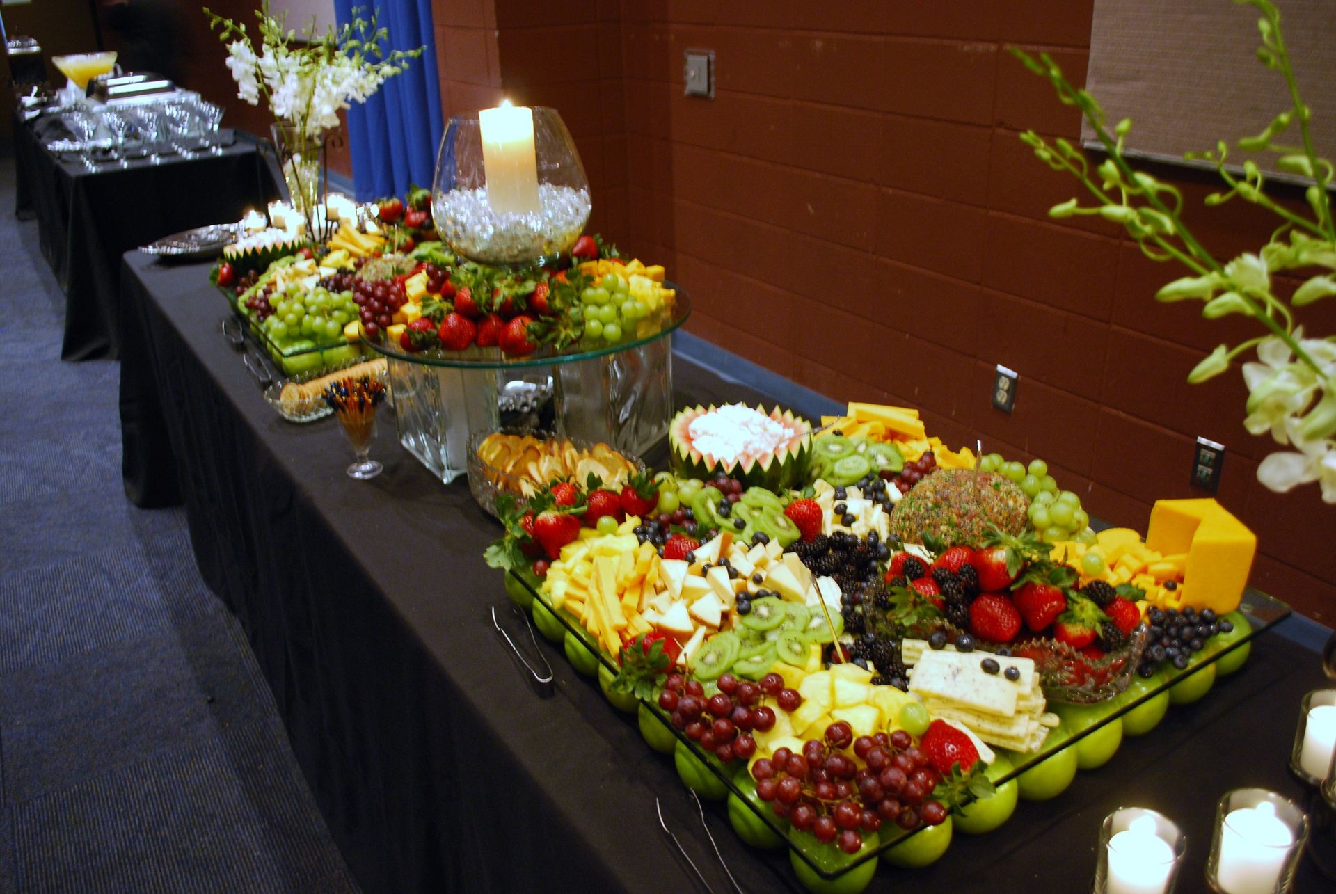 Buffet table with trays of fresh fruit and cheese on a black tablecloth against a brick wall