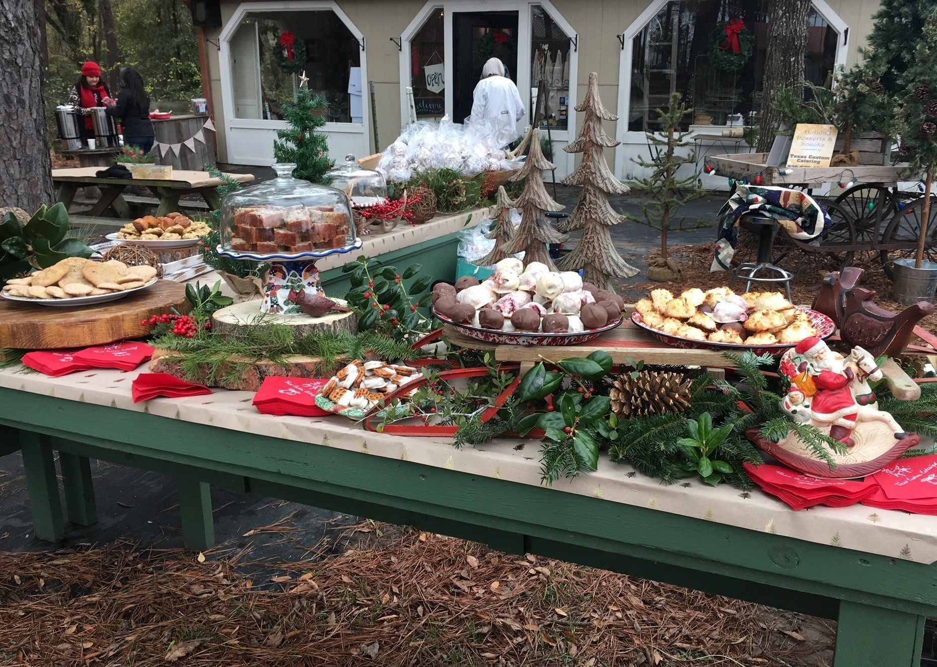 Outdoor holiday buffet table covered with festive food, pine branches, and red decorations in a courtyard