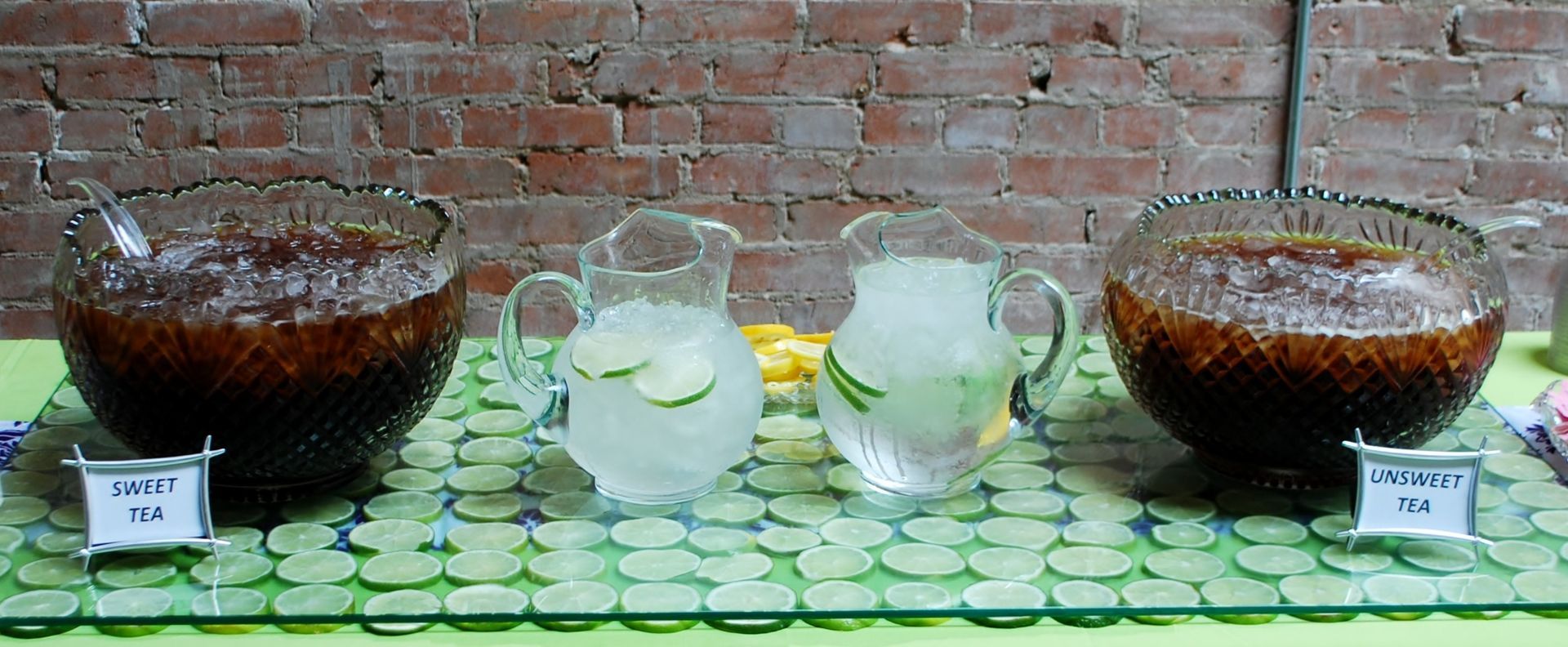 Glass pitchers and bowls of dark and light drinks displayed on a tiled table against a brick wall