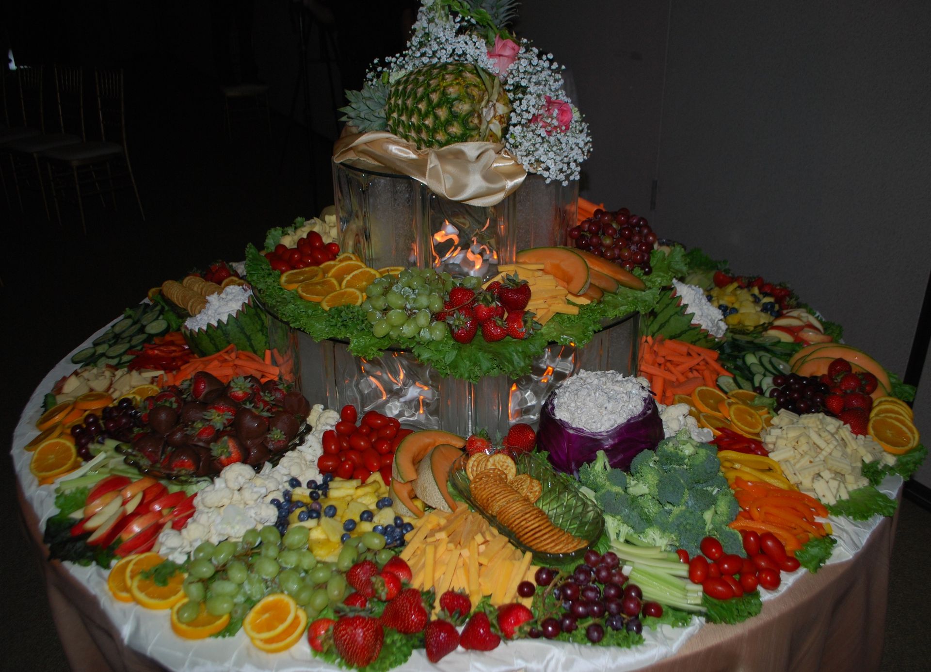 Large circular buffet table with assorted fruits, vegetables, bread, and flowers arranged decoratively on white cloth