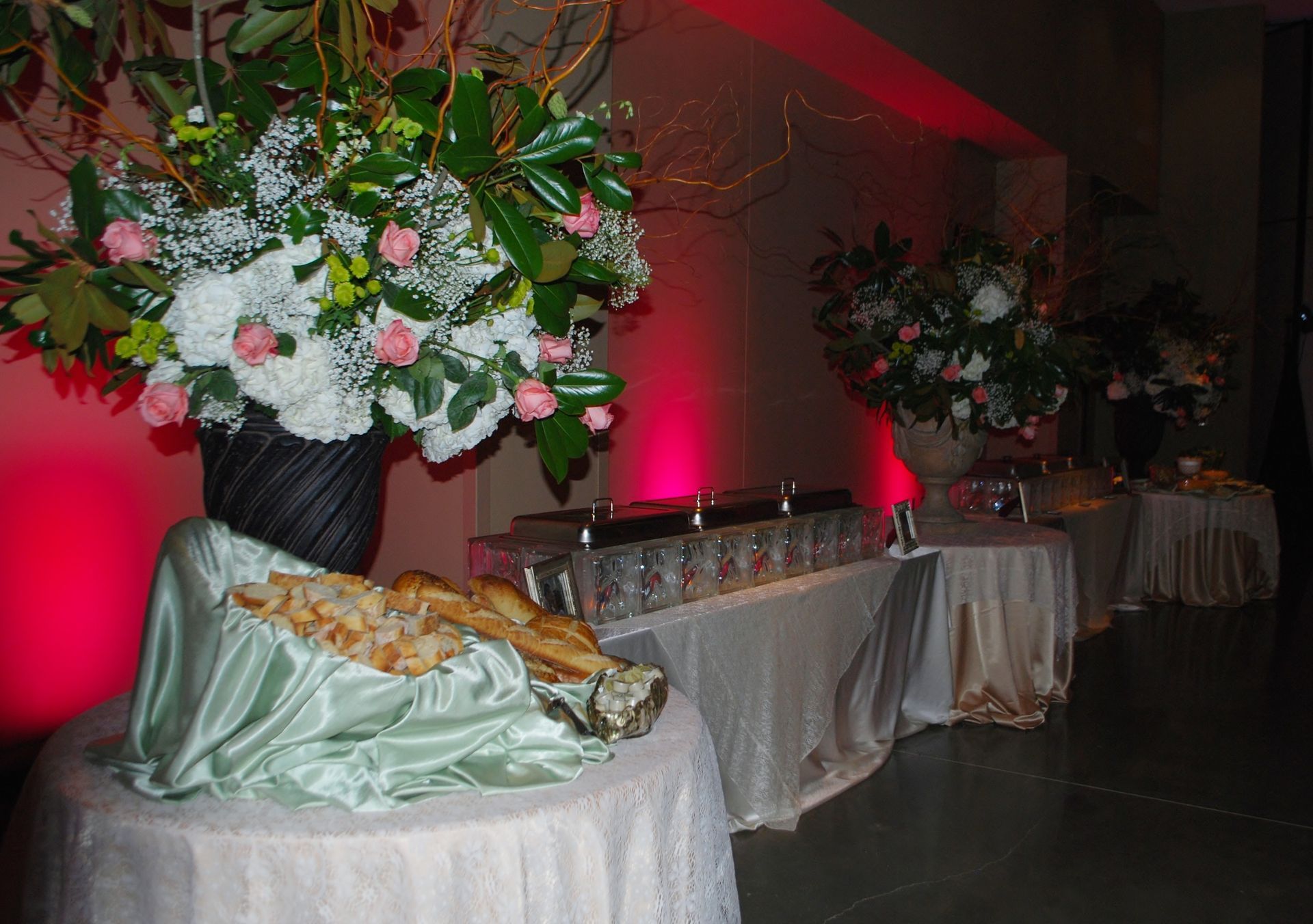 Decorated banquet table with floral arrangements, pink uplighting, and a covered dish at an event venue