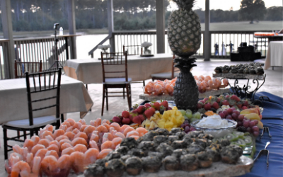 Outdoor buffet table with assorted fruit and dessert trays by a waterfront railing
