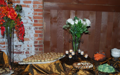 Decorated buffet table with gold cloth, white flowers, candles, and food trays against a brick wall