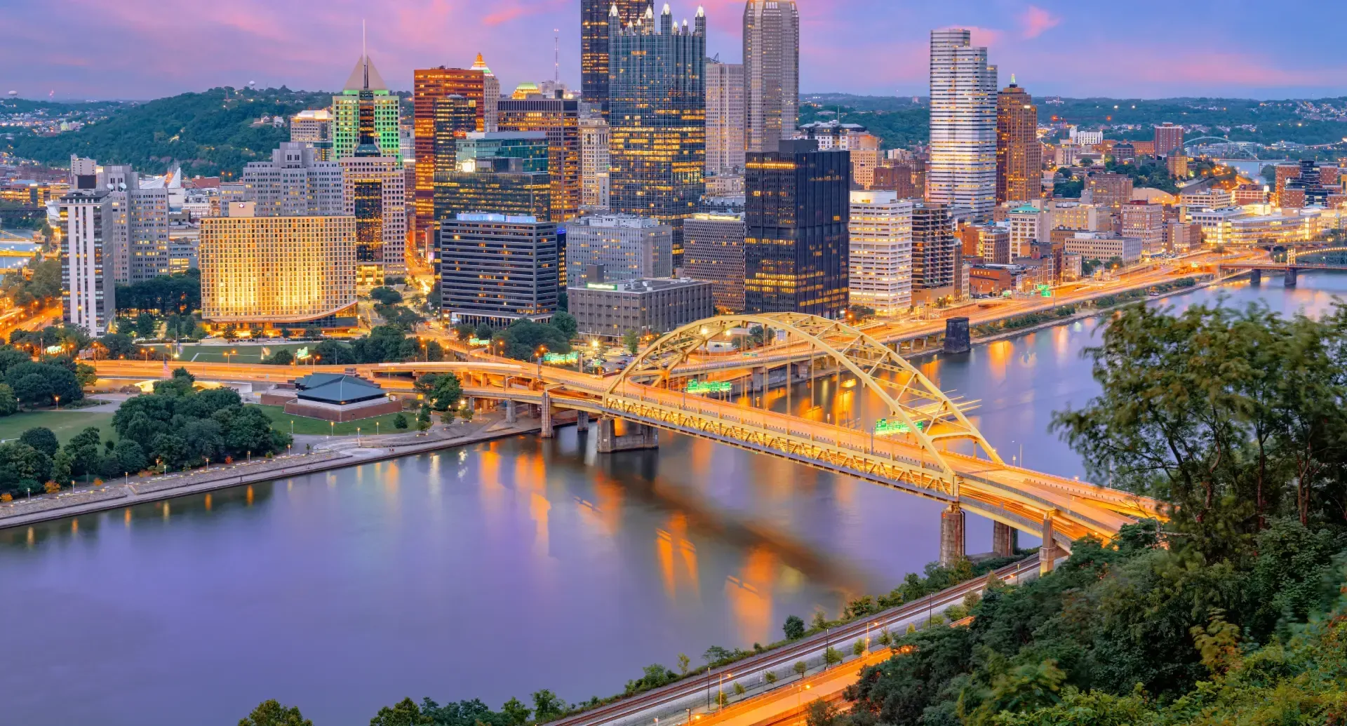 City skyline at dusk with a lit bridge over a river and reflections on the water