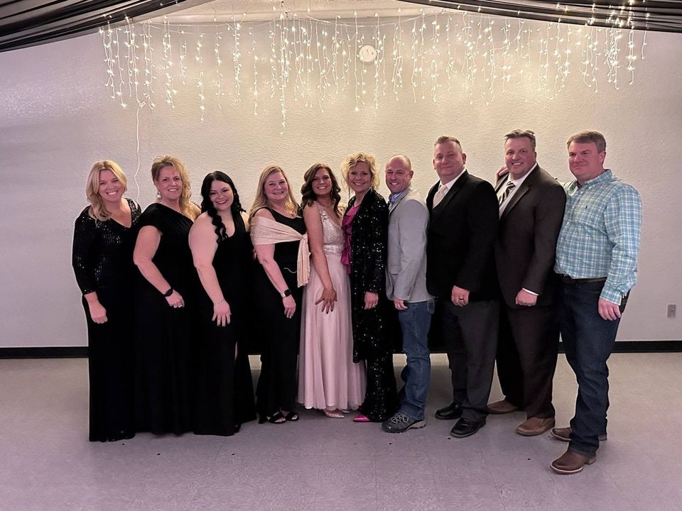Group of eleven people posing for photo, formal attire in a brightly lit room with string lights.