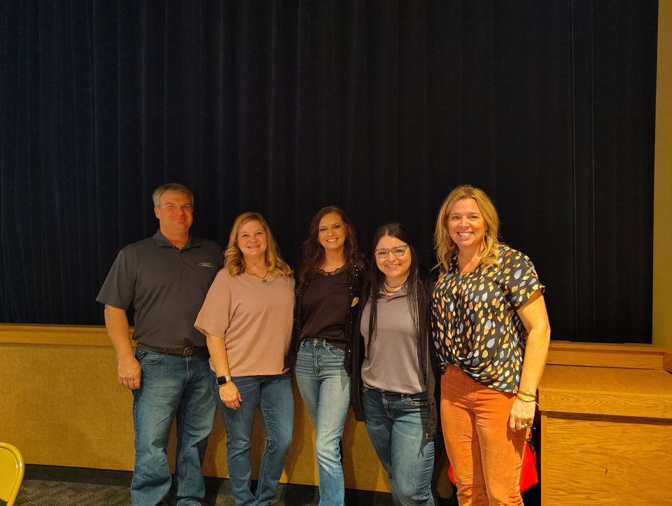 Five people stand in front of a dark curtain. Two women are on either end. The others are wearing jeans.