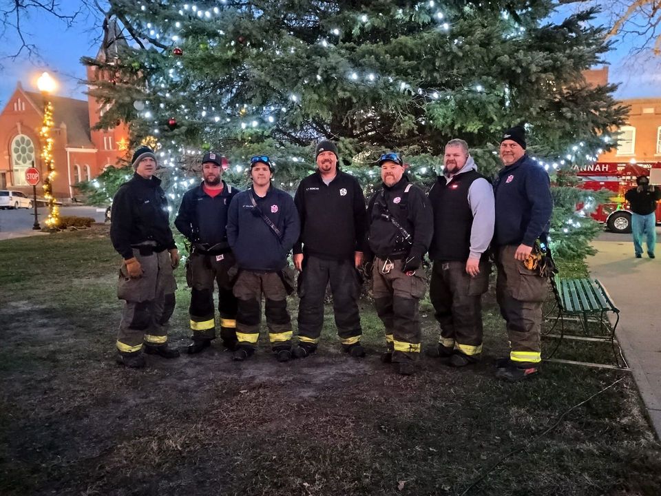 Firefighters pose in front of a lit Christmas tree. They wear uniforms and stand outside, near a building and fire truck.