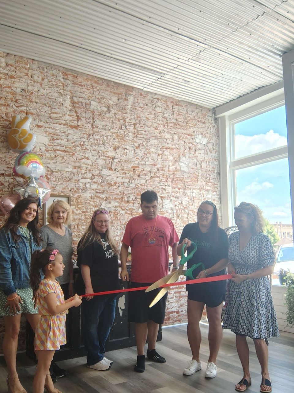 People cutting a red ribbon with large scissors at a business opening. Balloons and brick wall in the background.