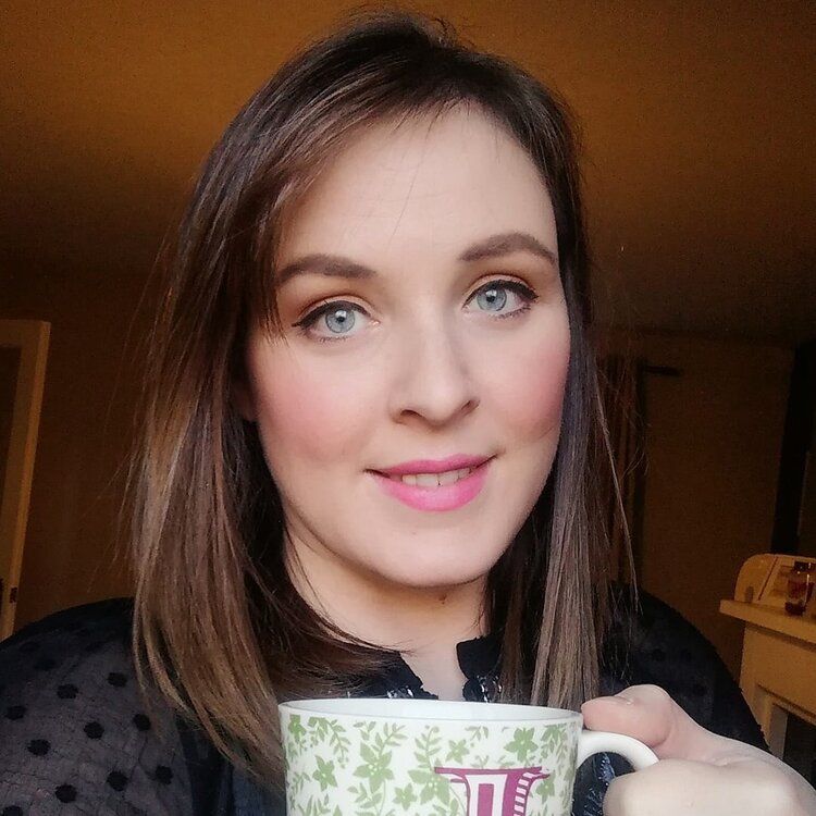 Woman with light skin, blue eyes, and pink lipstick holding a floral mug, smiling.