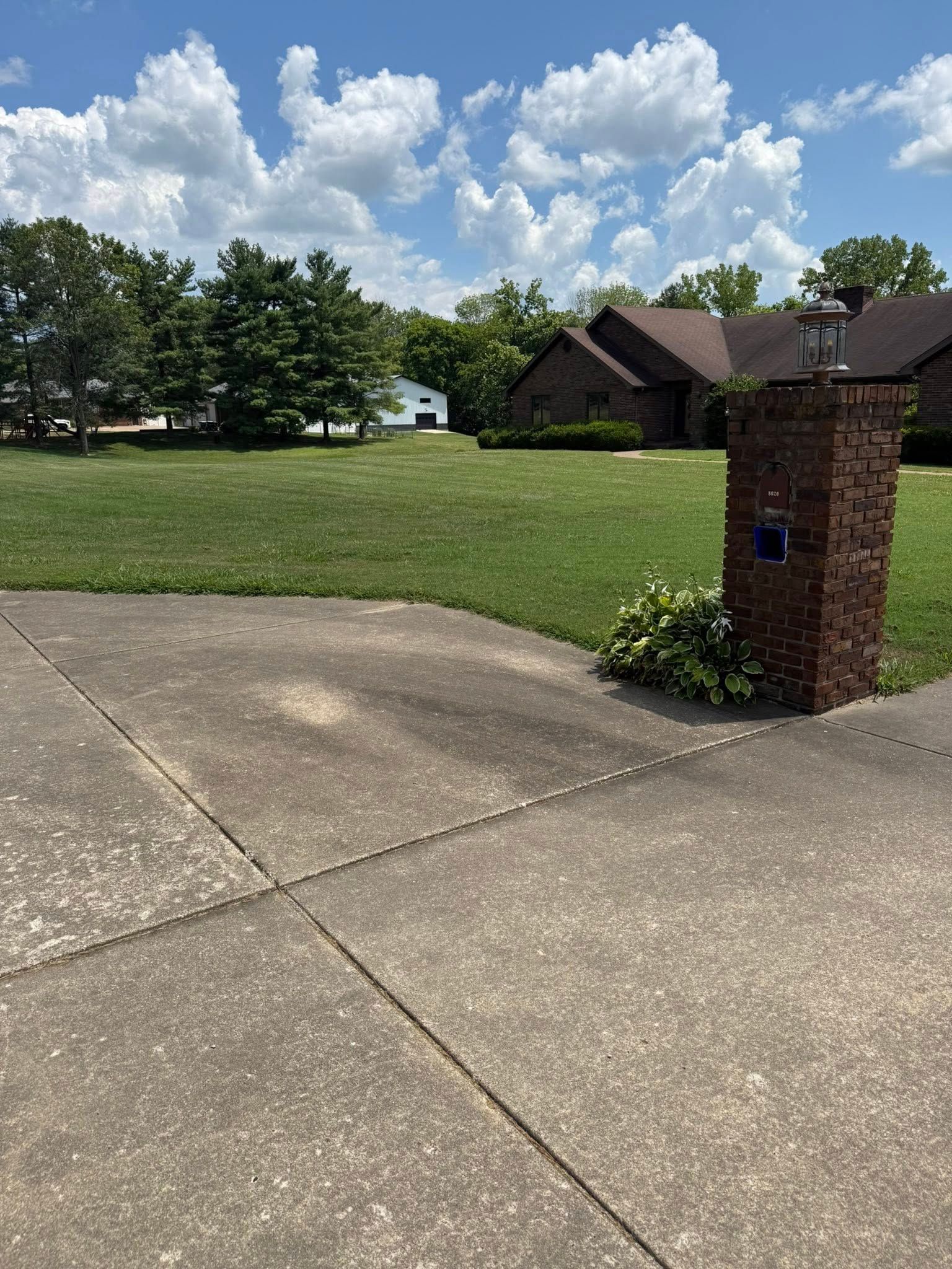 A brick pillar with a mailbox stands on a concrete driveway with a grassy yard, trees, and a house in the background.