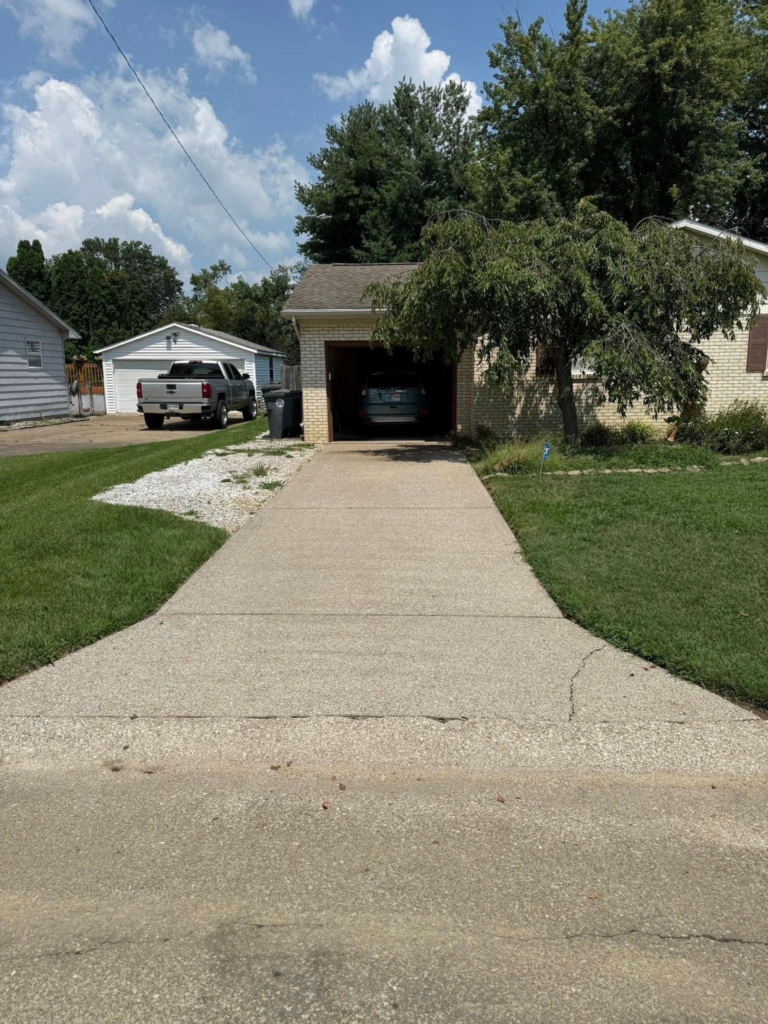 Concrete driveway leading to a garage with a car inside; green lawn surrounds the driveway, a truck is parked in the driveway across from the garage.
