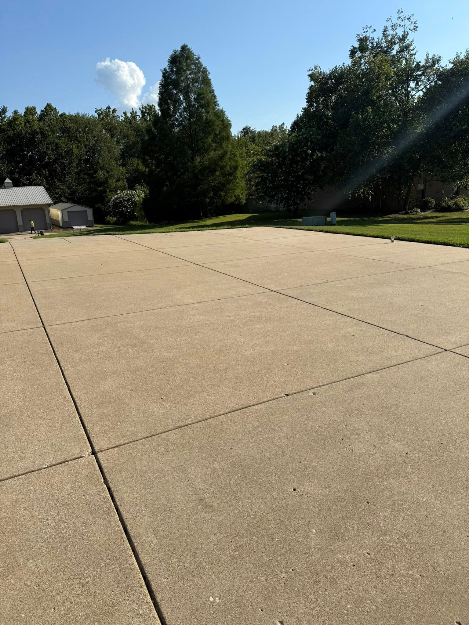 Large concrete patio with separation lines. Trees and a small building in the background under a blue sky.