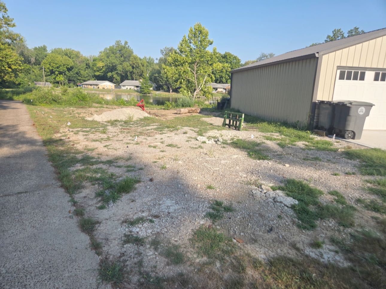 Gravel yard next to a driveway and beige building with trash bins. Trees and houses in the background under a blue sky.