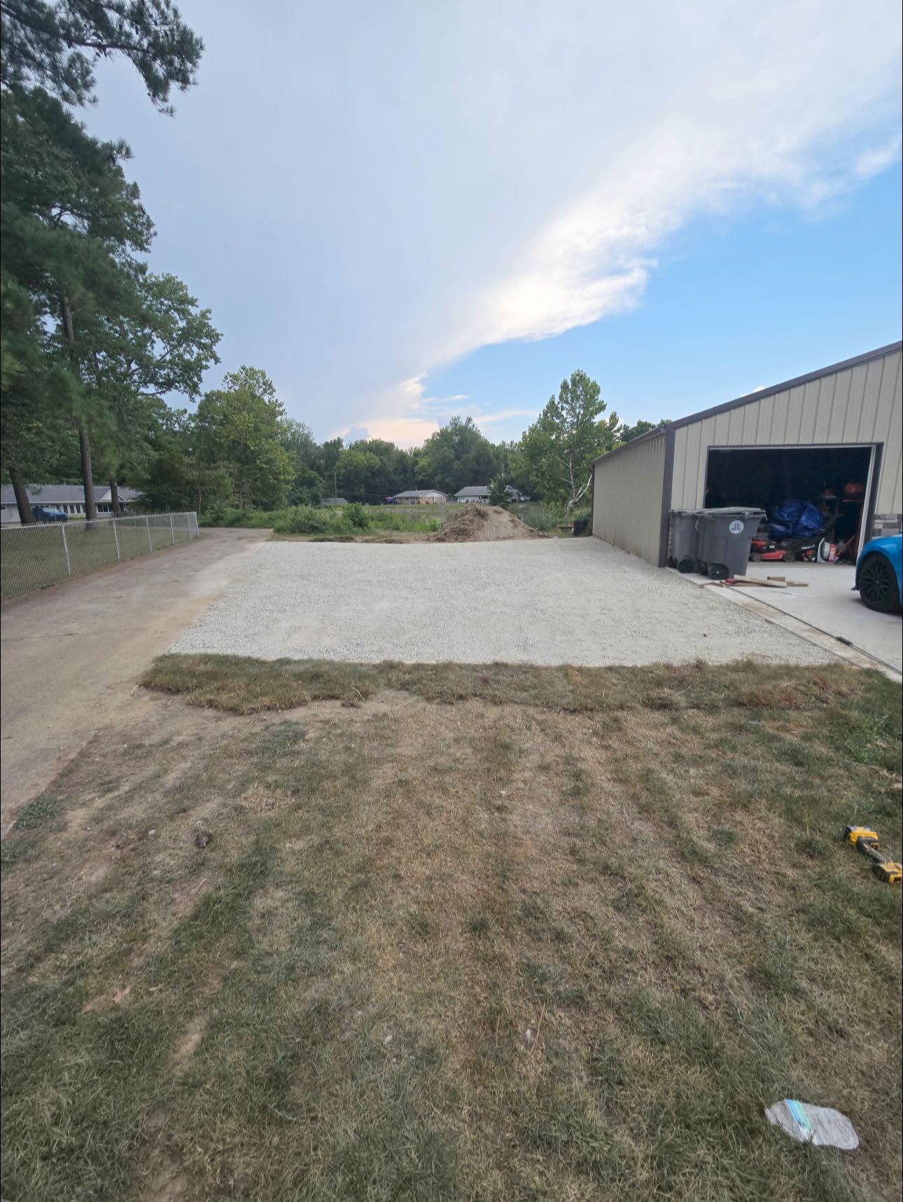 Gravel lot bordered by dry grass, next to a garage and driveway. Trees and sky in the background.